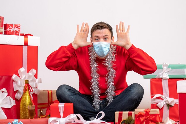 Front view of young man in mask sitting around xmas presents on white wall