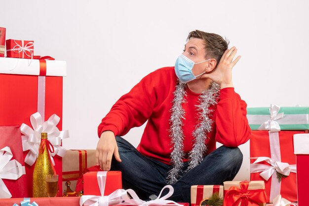 Front view of young man in mask sitting around xmas presents on white wall