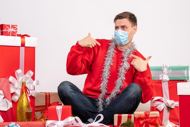 Front view of young man in mask sitting around xmas presents on white wall