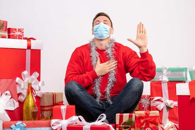 Front view of young man in mask sitting around xmas presents on white wall