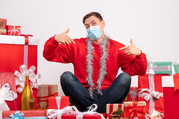 Вид спереди молодого человека в маске, сидящего вокруг рождественских подарков на белой стене