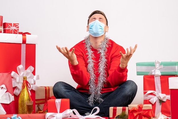 Front view of young man in mask sitting around xmas presents on white wall