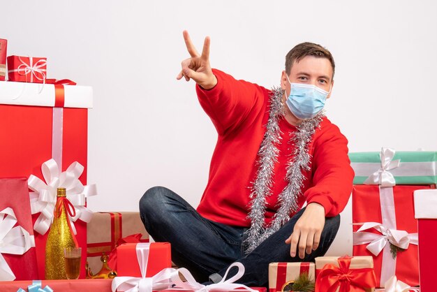 Front view of young man in mask sitting around xmas presents on white wall