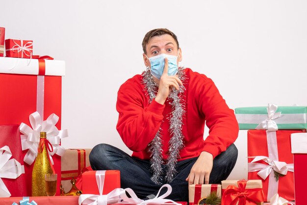 Front view of young man in mask sitting around xmas presents on white wall