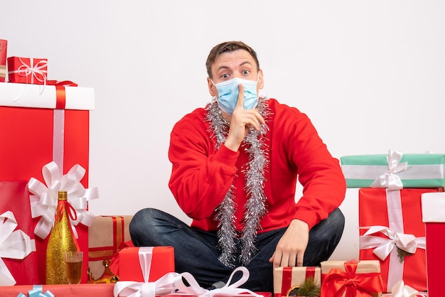 Front view of young man in mask sitting around xmas presents on white wall