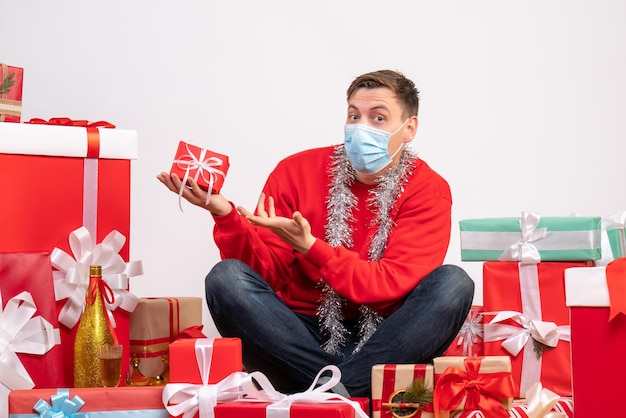 Front view of young man in mask sitting around xmas presents on white wall