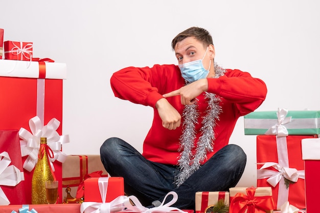 Front view of young man in mask sitting around xmas presents on white wall