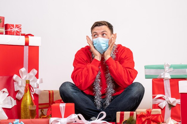 Front view of young man in mask sitting around xmas presents on white wall