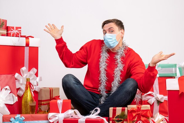 Front view of young man in mask sitting around xmas presents on white wall