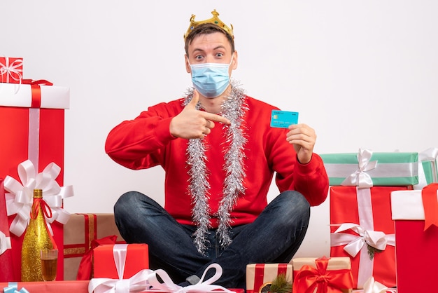 Vista frontale del giovane in maschera seduto intorno ai regali di natale con carta bancaria sul muro bianco