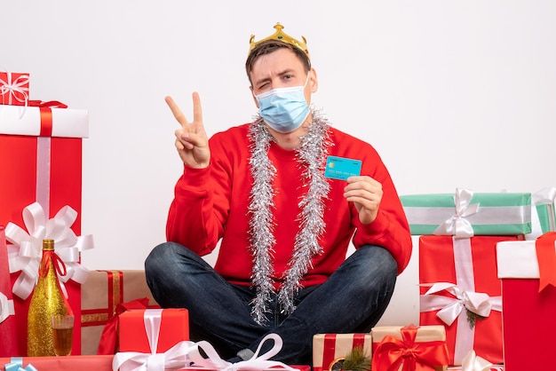 Вид спереди молодого человека в маске, сидящего вокруг рождественских подарков с банковской картой на белой стене