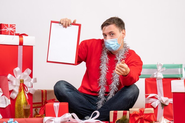 Front view of young man in mask sitting around xmas presents with note on the white wall