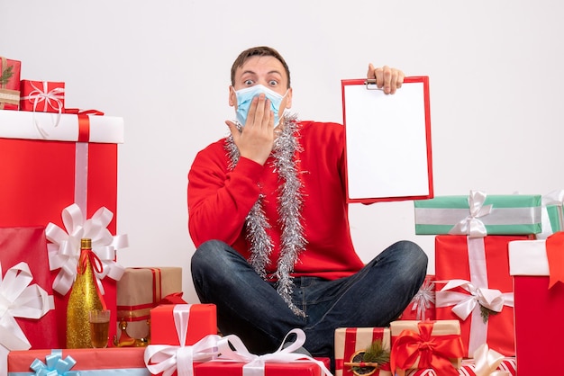 Front view of young man in mask sitting around xmas presents with note on white wall