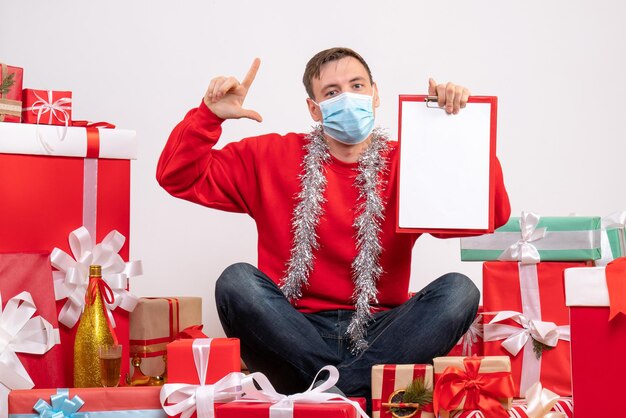 Front view of young man in mask sitting around xmas presents with note on white wall