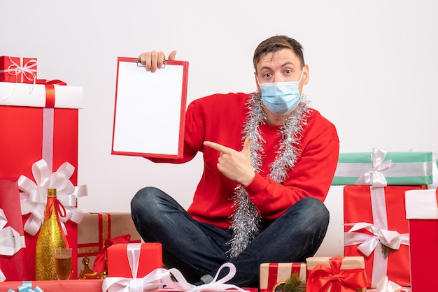 Front view of young man in mask sitting around xmas presents with note on white wall