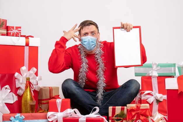 Front view of young man in mask sitting around xmas presents with note on white wall