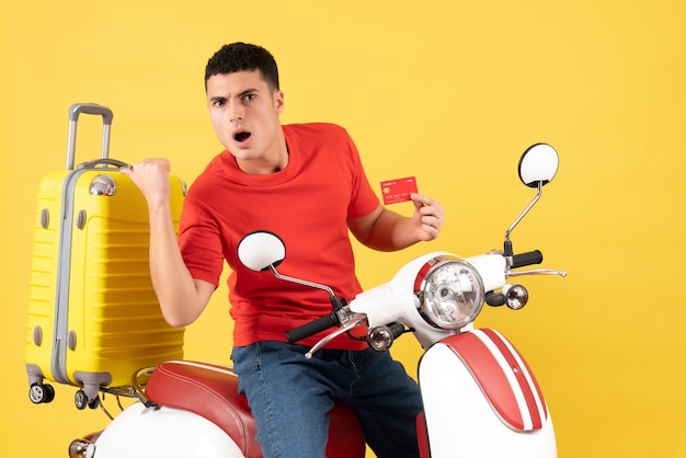 Front view young man on moped holding credit card pointing at his suitcase