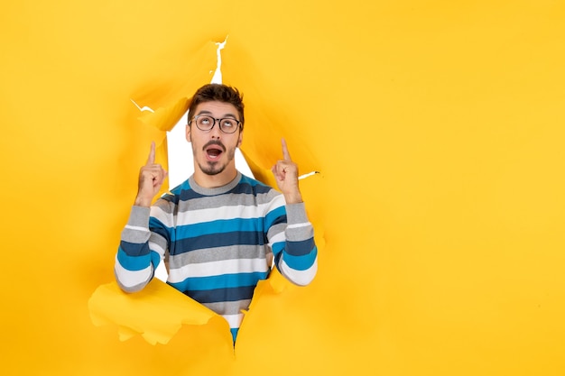 Front view young man pointing fingers up looking through ripped paper yellow wall