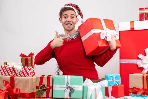 Front view young man pointing at giftbox sitting around xmas gifts