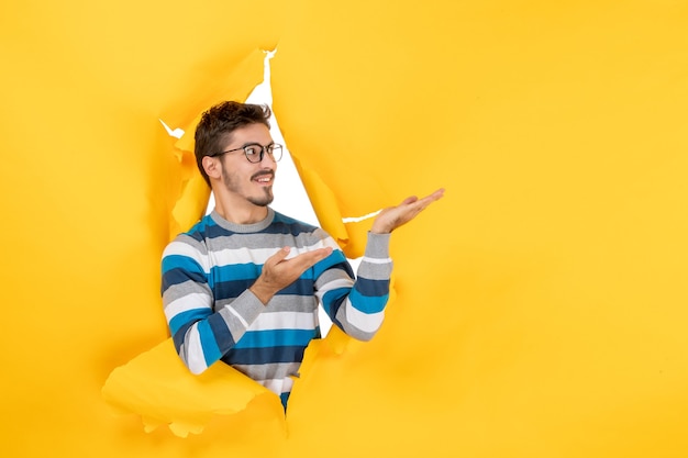 Free photo front view young man pointing at left peeping through hole in paper yellow wall