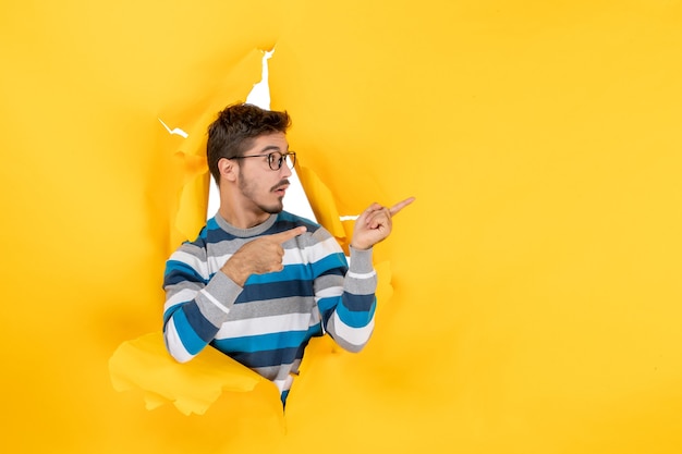 Front view young man pointing at right peeping through hole in paper yellow wall