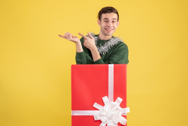 Front view young man pointing at something standing behind big giftbox on yellow 