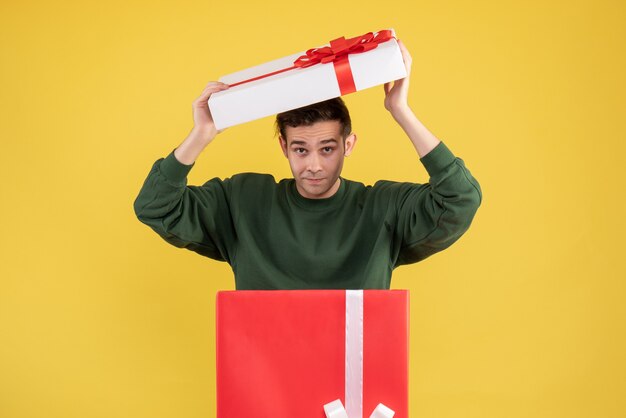 Front view young man putting cover over his head standing behind big giftbox on yellow 