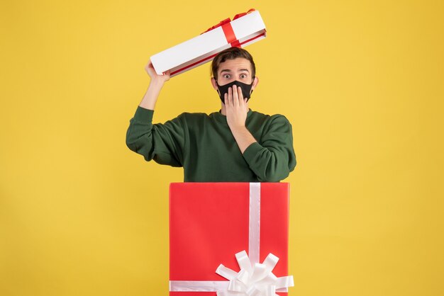 Front view young man putting cover over his head standing behind big giftbox on yellow 