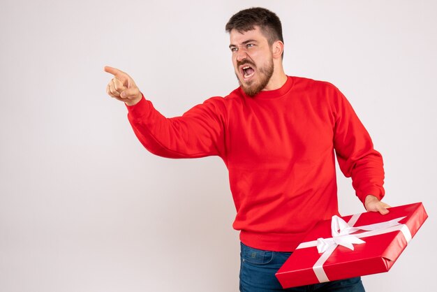 Front view of young man in red shirt holding christmas present and arguing with someone on white wall