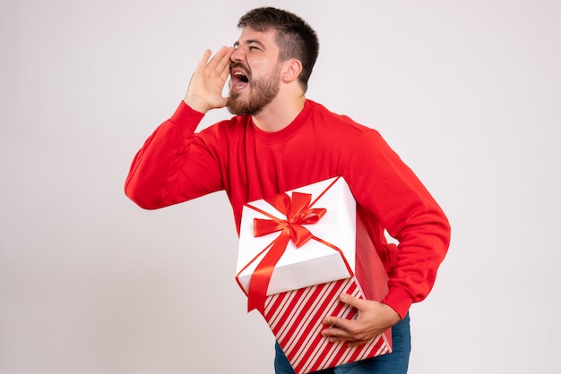 상자에 크리스마스 선물을 들고 흰 벽에 비명을 지르는 빨간 셔츠에있는 젊은 남자의 전면보기