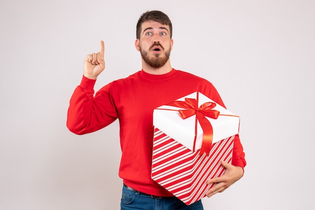 Front view of young man in red shirt holding christmas present in box on a white wall