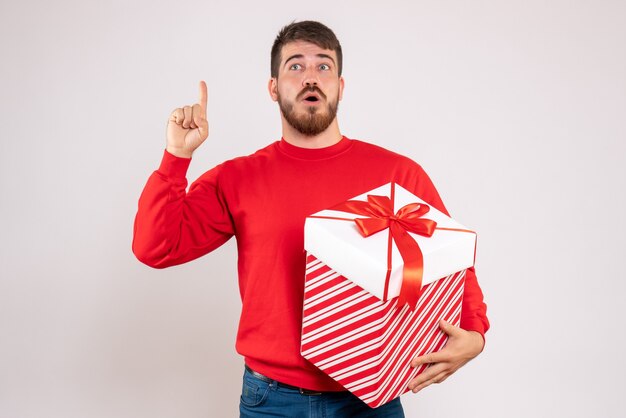 Front view of young man in red shirt holding christmas present in box on a white wall