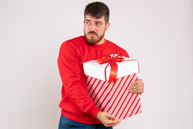 Front view of young man in red shirt holding christmas present in box on a white wall