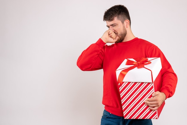 Front view of young man in red shirt holding christmas present in box on white wall