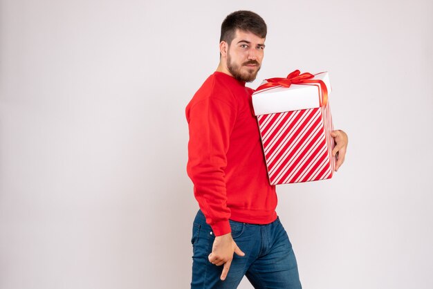 Front view of young man in red shirt holding christmas present in box on white wall