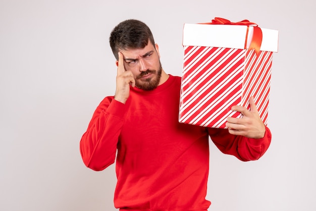 Вид спереди молодого человека в красной рубашке, держащего рождественский подарок в коробке на белой стене