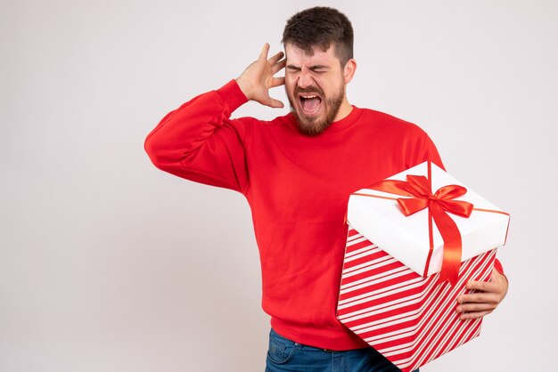 Front view of young man in red shirt holding christmas present in box on white wall