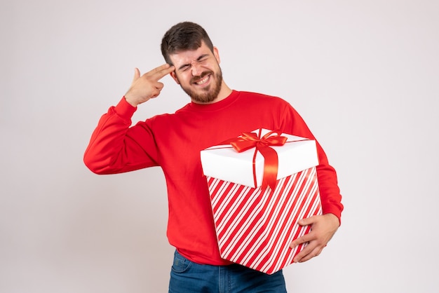Вид спереди молодого человека в красной рубашке, держащего рождественский подарок в коробке на белой стене