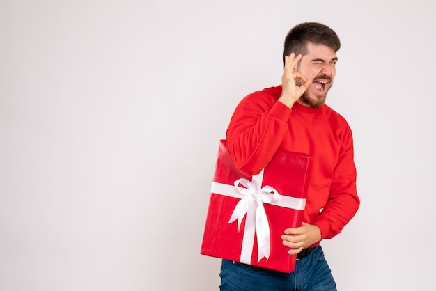 白い壁にクリスマスプレゼントを保持している赤いシャツを着た若い男の正面図