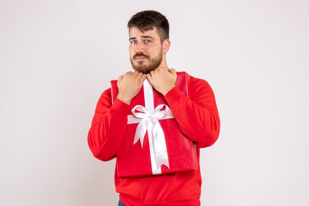 Front view of young man in red shirt holding christmas present on white wall