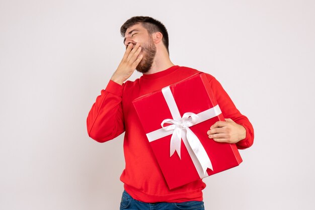 Front view of young man in red shirt holding christmas present and yawning on white wall