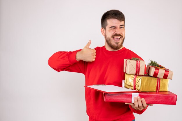 Front view of young man in red shirt holding christmas presents arguing on white wall