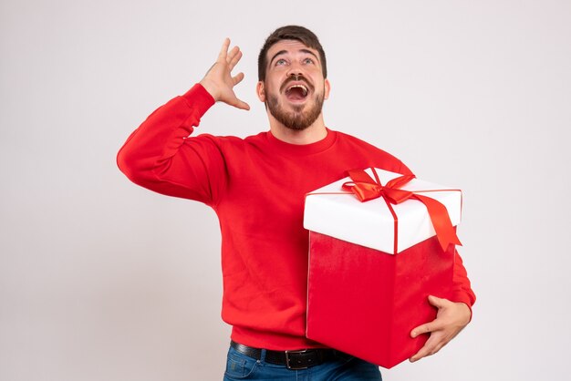 Front view of young man in red shirt holding xmas present in box on white wall