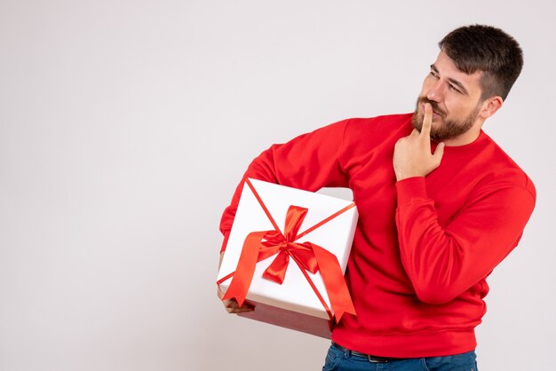 Front view of young man in red shirt holding xmas present in box on white wall