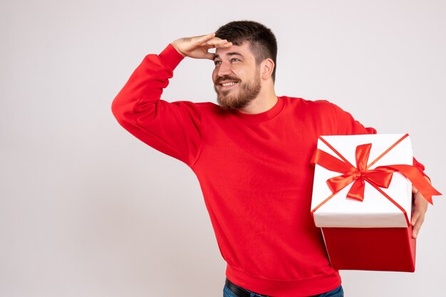 Front view of young man in red shirt holding xmas present in box on white wall