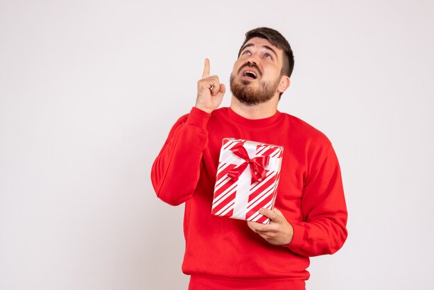 Front view of young man in red shirt holding xmas present on a white wall