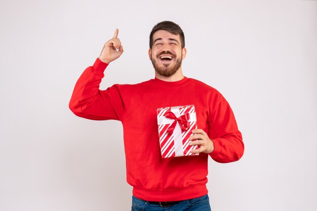 Вид спереди молодого человека в красной рубашке, держащего рождественский подарок на белой стене