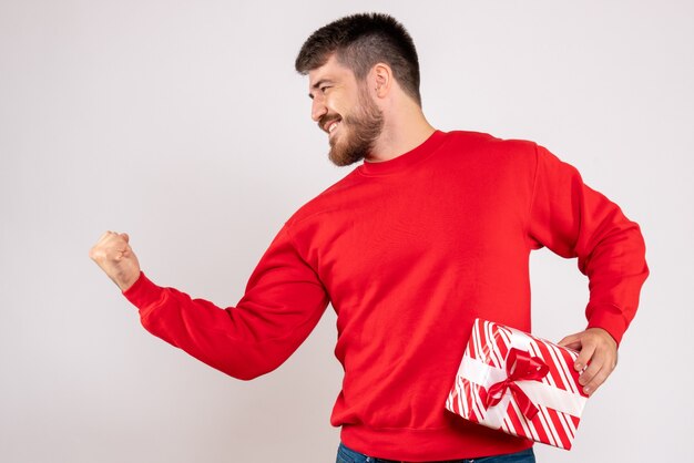 白い壁にクリスマスプレゼントを保持している赤いシャツを着た若い男の正面図