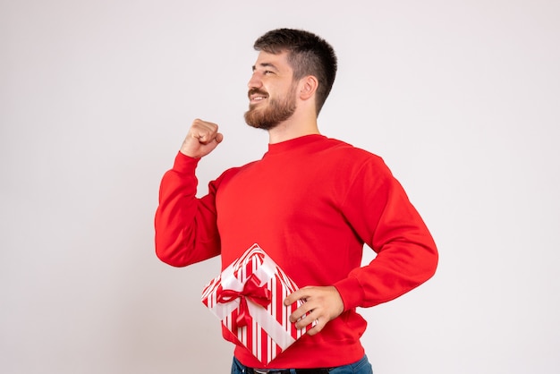 Front view of young man in red shirt holding xmas present on white wall