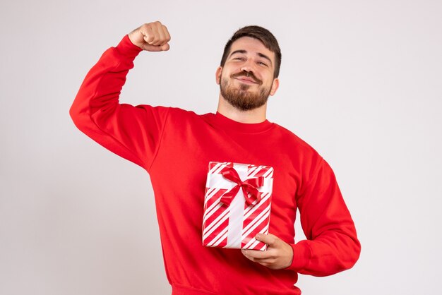 Front view of young man in red shirt holding xmas present on white wall